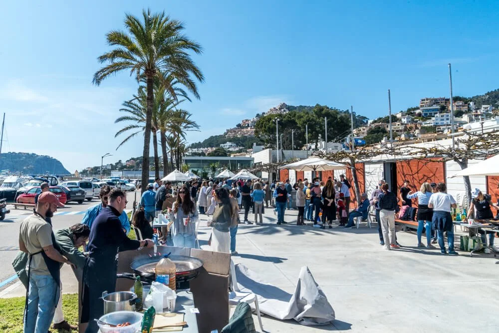 Evento al aire libre con muchas personas, vendedores y mesas con comida, junto a un puerto con barcos y palmeras.