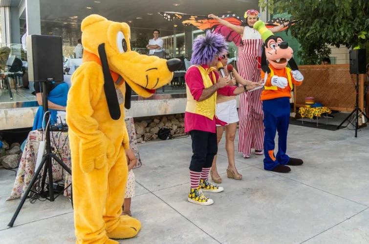 Personajes disfrazados, incluyendo a Pluto y Goofy, en un evento al aire libre con niños y payasos, realizando una actuación o show para niños.