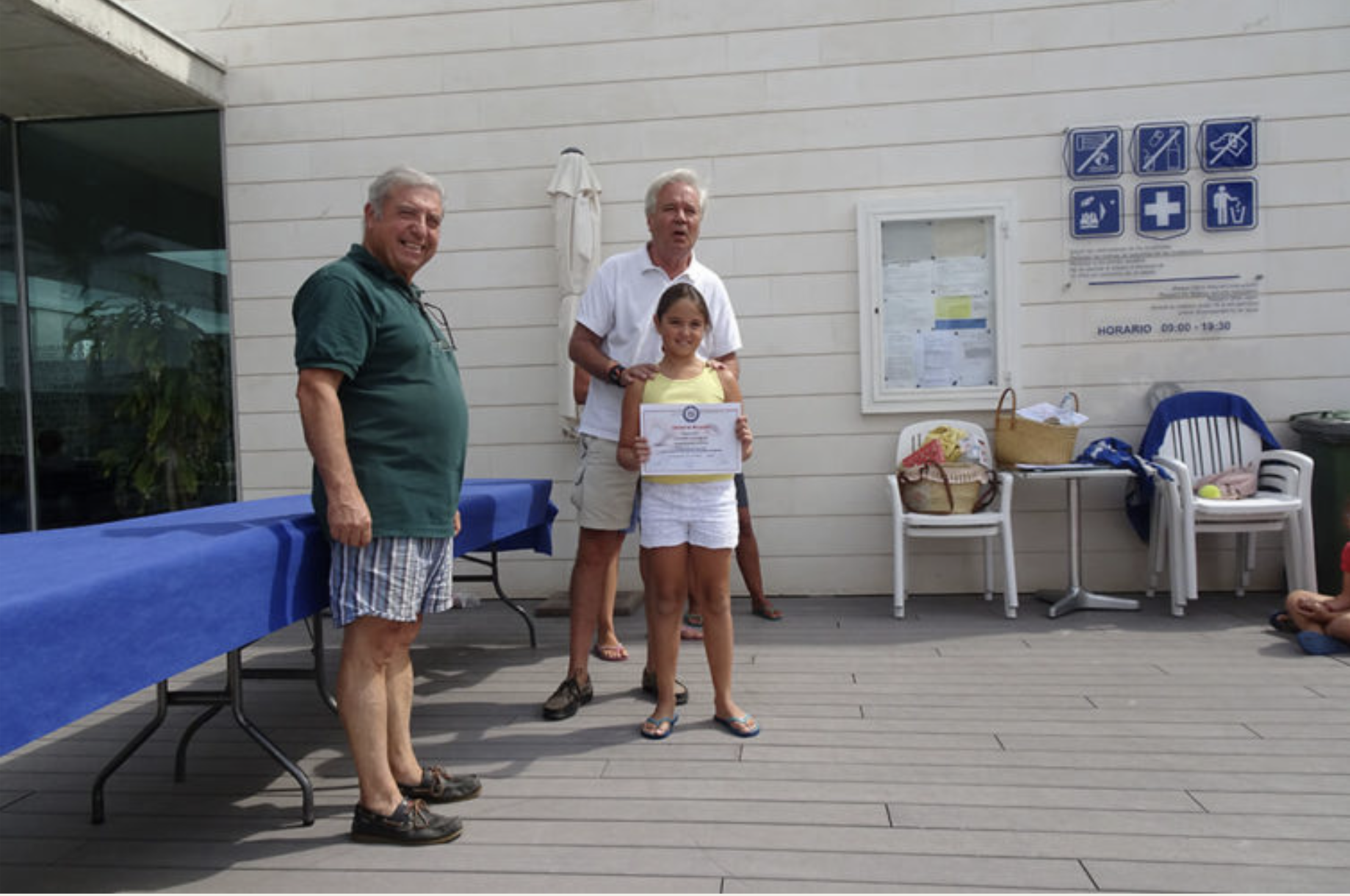 Un grupo de personas, incluyendo una niña recibiendo un certificado, en un evento al aire libre con mesas y sillas. Dos hombres y una mujer en el fondo, todos sonriendo.