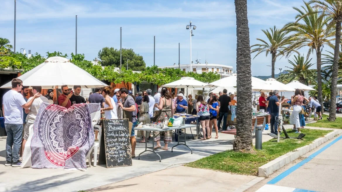 Ferias al aire libre con puestos de comida y personas disfrutando bajo sombrillas y palmeras en un día soleado.