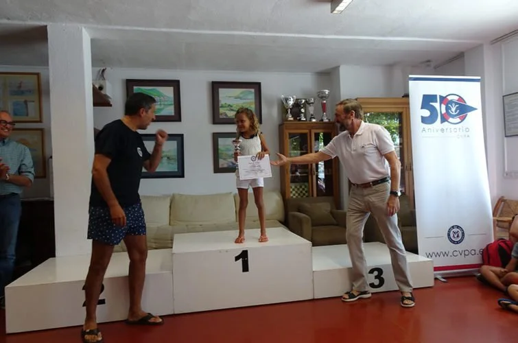 Niña en un podio de primer lugar recibiendo un premio en una ceremonia, con un adulto entregándole el premio y otros asistentes observando, en un salón decorado con trofeos y cuadros.