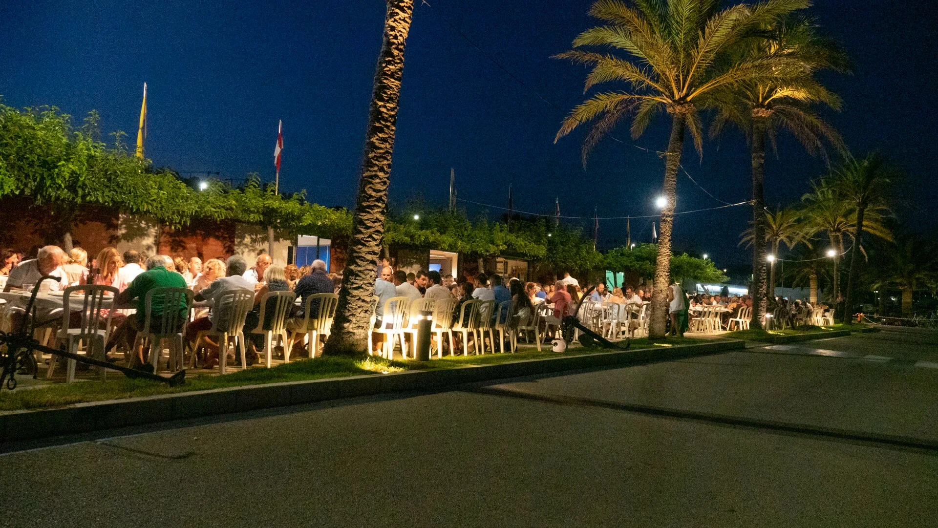 Reunión de muchas personas cenando al aire libre por la noche, rodeadas de palmeras y árboles, con luces decorativas y banderas en el fondo.