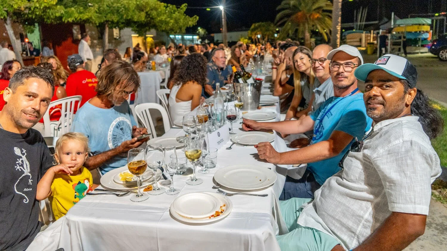 Grupo de personas disfrutando de una comida en una celebración al aire libre por la noche, con una fila larga de mesas decoradas con vasos, platos y botellas de vino.