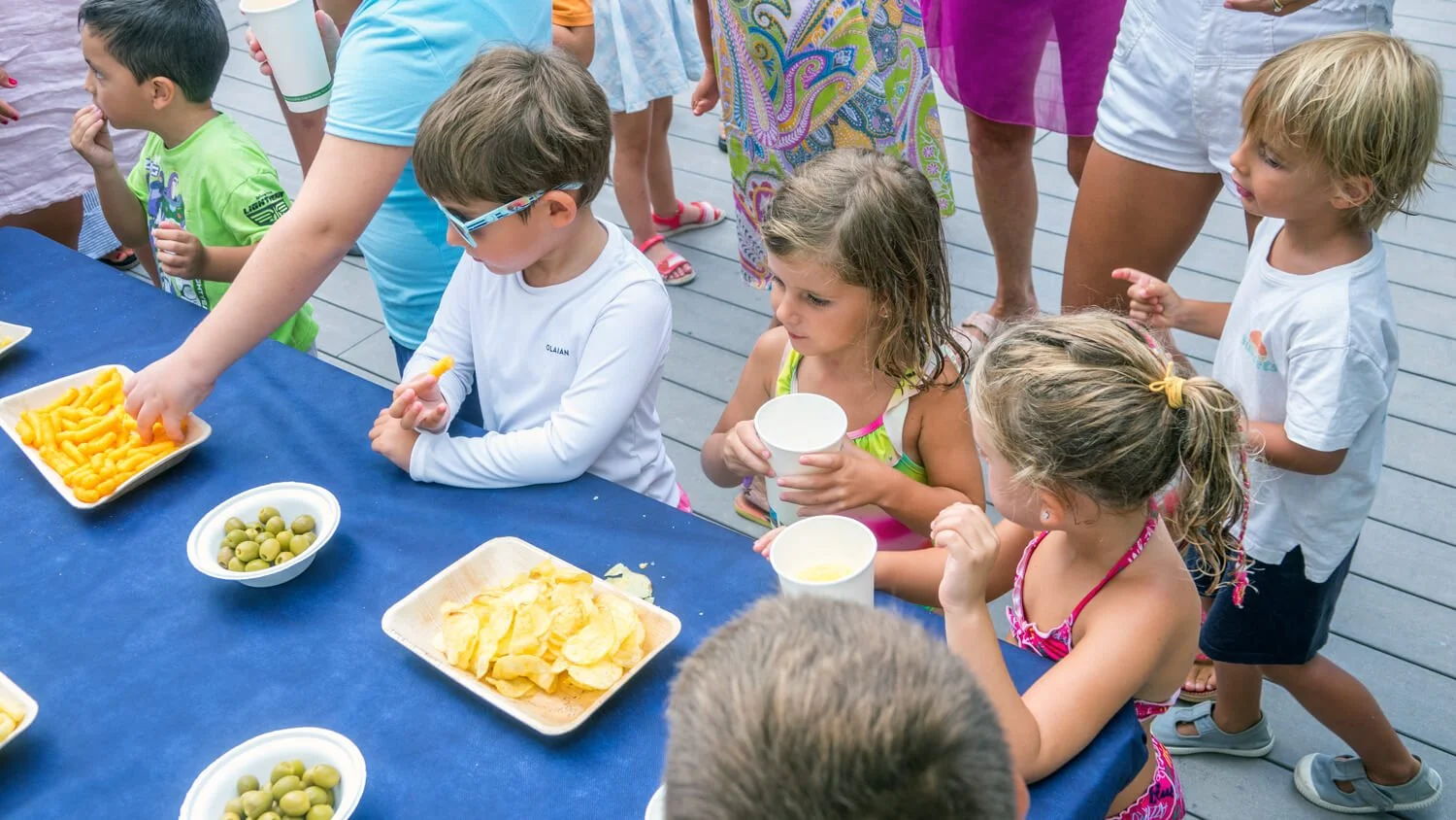 Niños en línea con adultos en una fiesta infantil, sirviéndose snacks como patatas fritas, uvas y bebidas en una mesa con mantel azul en un espacio al aire libre.