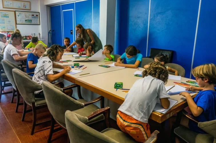 Niños en clase de arte, algunos pintando y otros escuchando a la maestra en un aula.