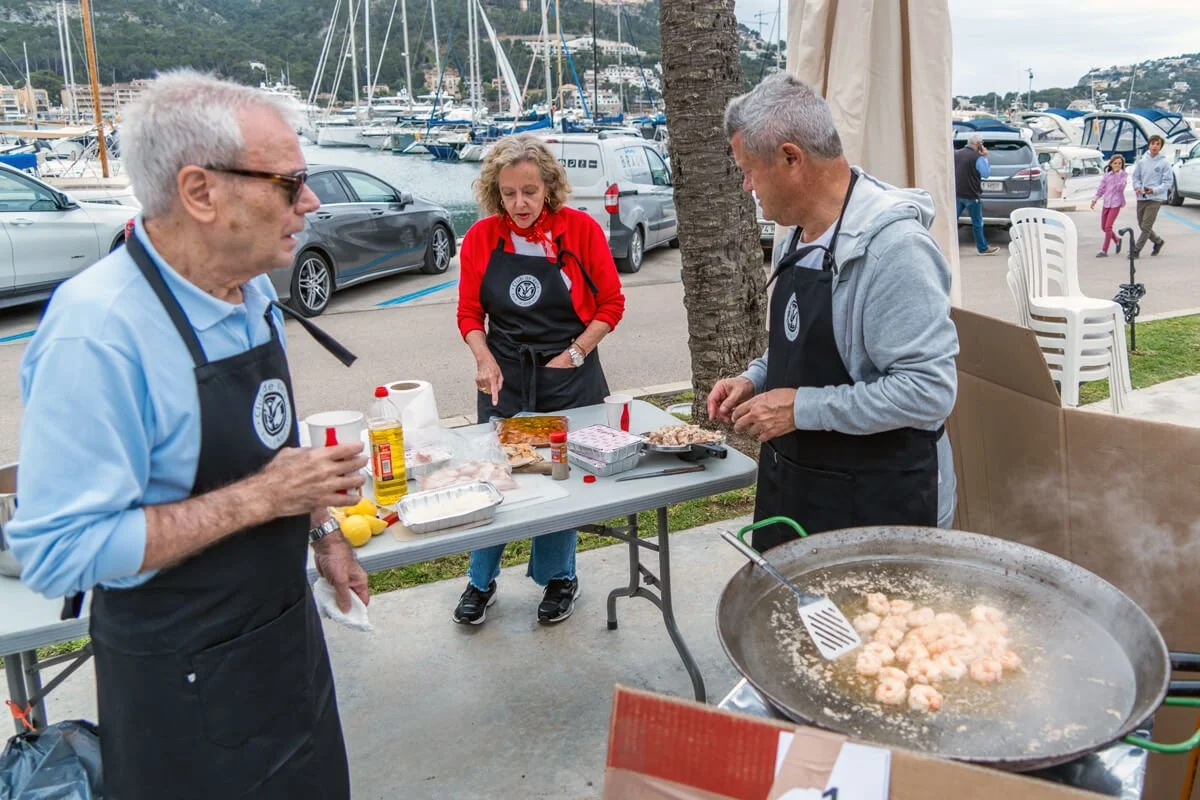 tres personas cocinando mariscos en una comida al aire libre cerca del puerto, con barcos y autos de fondo