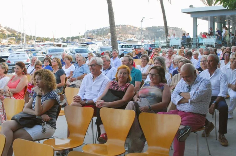 Personas asistiendo a un evento al aire libre, sentadas en sillas en una terraza con vista al mar y yates.