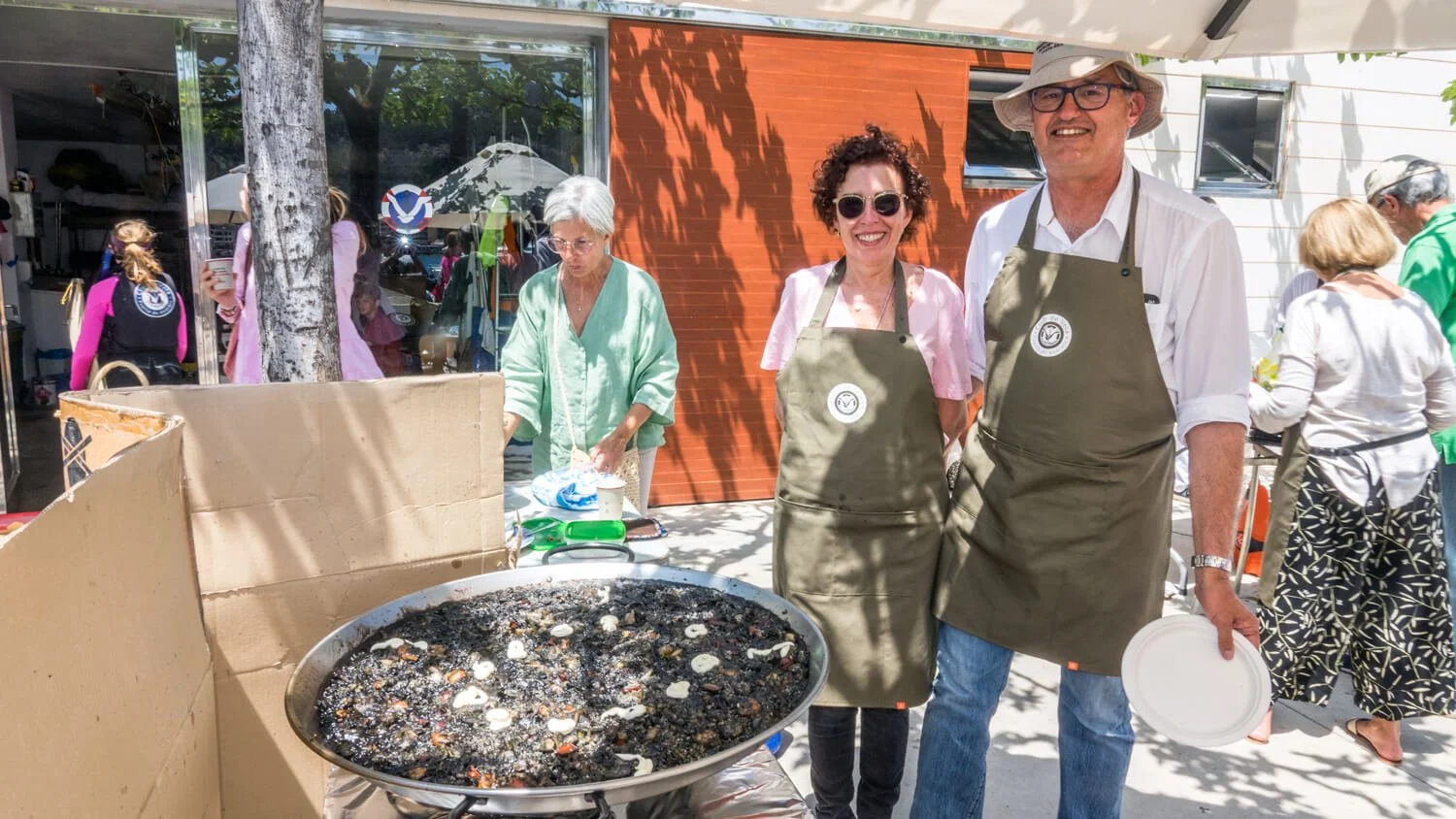 Personas cocinando y sirviendo comida en un evento al aire libre, con un grupo en el fondo y un gran sartén con comida en primer plano.