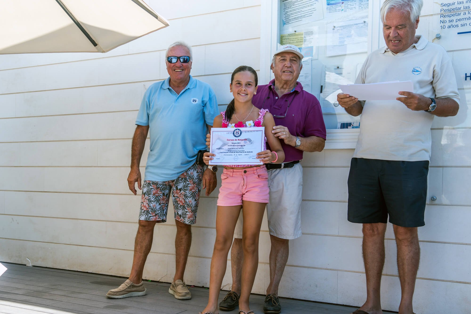 Cuatro personas, un joven y tres adultos mayores, posan con una niña sosteniendo un certificado, en un ambiente de competencia o celebración deportiva, con carteles en la pared y un paraguas en el fondo.
