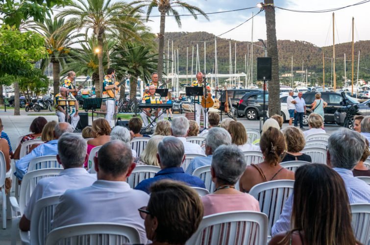 Un grupo de músicos tocando instrumentos en un escenario al aire libre junto a un muelle con yates, mientras una audiencia de personas observa sentada en sillas plásticas.