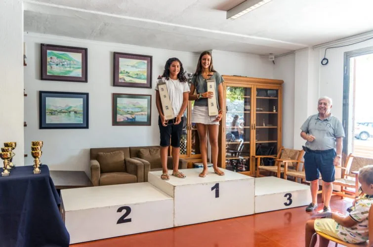 Dos niñas en el podio de premios, la mayor en primer lugar y la otra en segundo, sosteniendo trofeos, mientras una persona adulta observa y un niño mira sentado a la derecha.