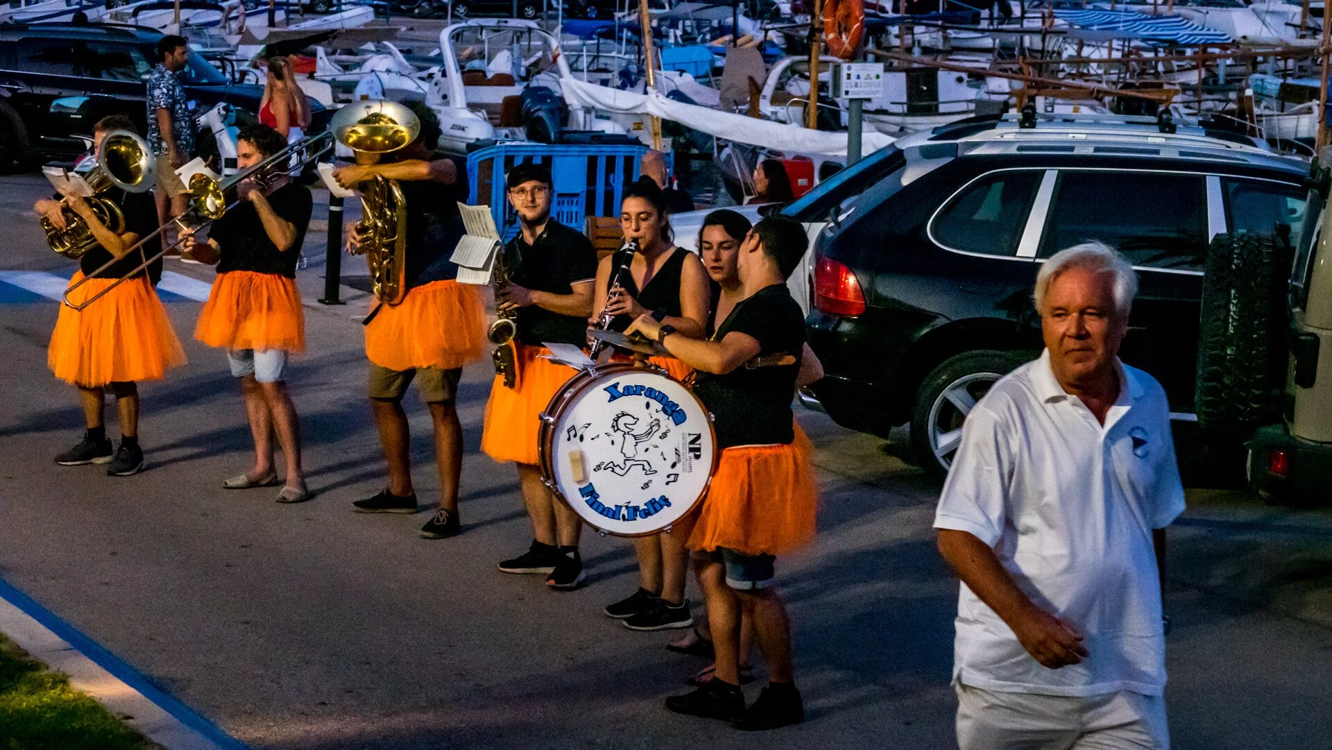 Grupo de músicos tocando instrumentos en una calle, con caritas y vinchas anaranjadas, durante el atardecer, cerca de vehículos estacionados y un muelle con embarcaciones.