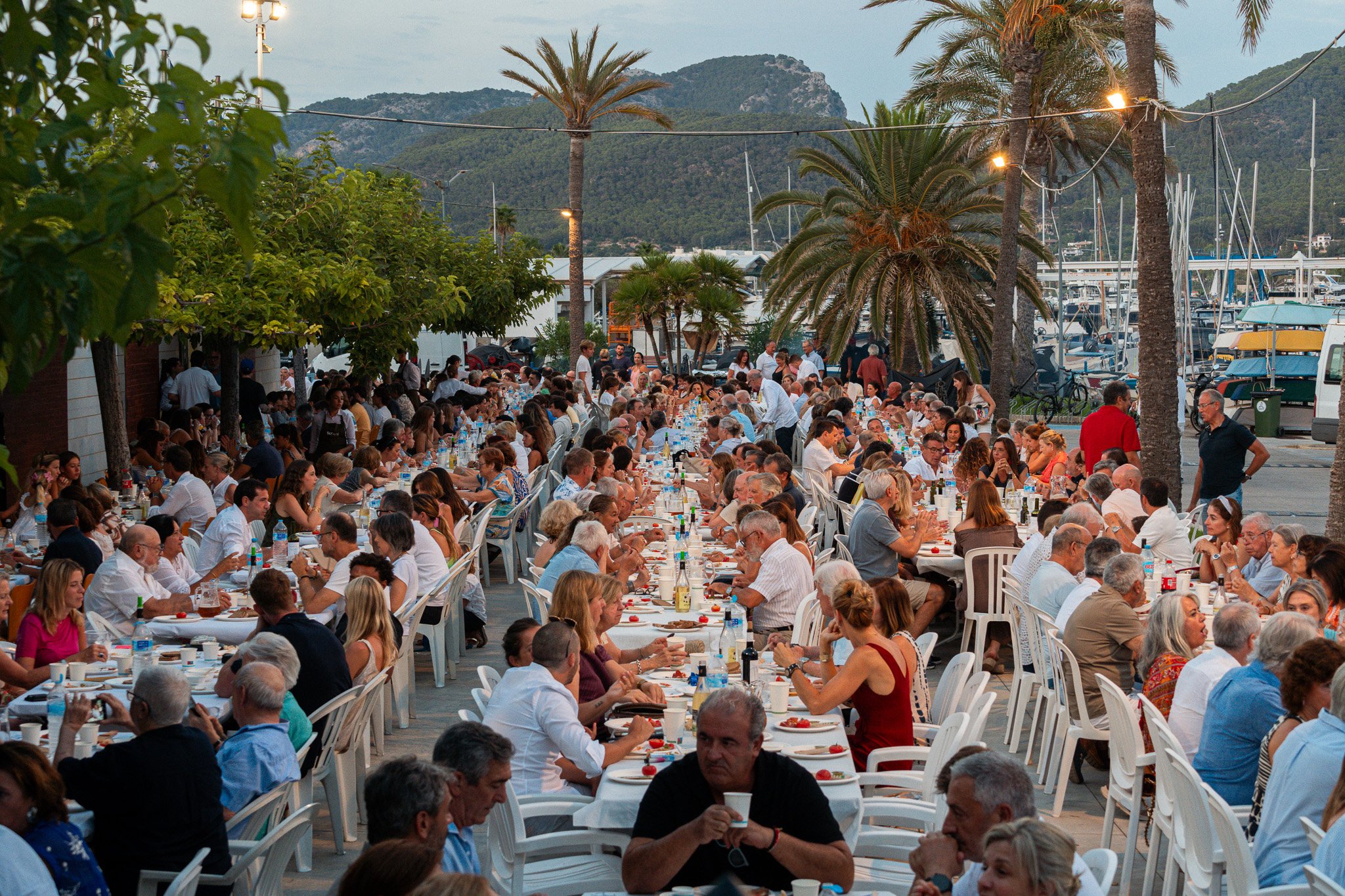 Una reunión o evento social al aire libre con muchas personas sentadas en mesas con comida y bebida, rodeadas de palmeras y cercanía a un puerto con barcos y yates, montañas al fondo y luces decorativas colgadas.