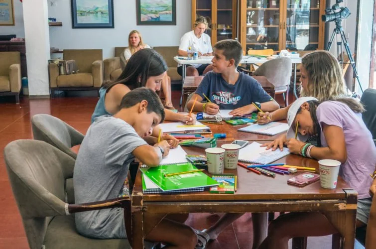 Niños y jóvenes estudiando y dibujando en una mesa en una sala que parece un aula o espacio de actividades, con adultos en el fondo y equipo de grabación.