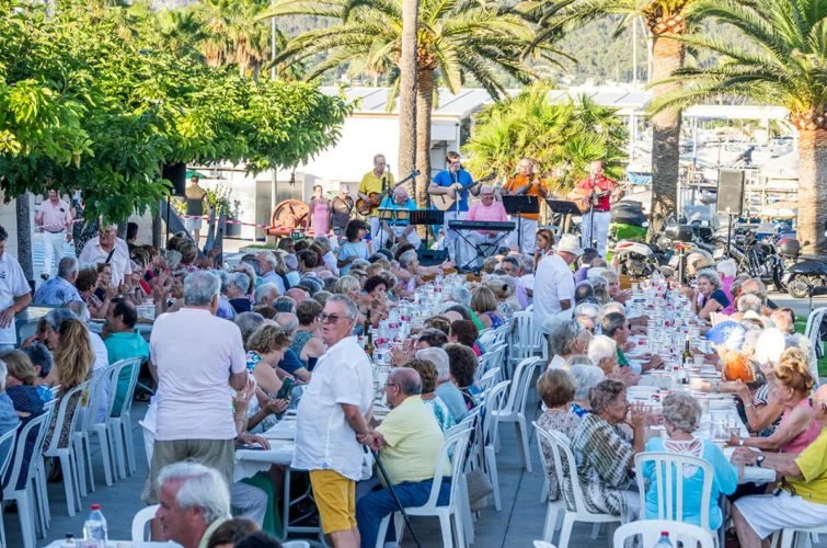 Concierto al aire libre con músicos tocando en el escenario y una gran multitud de personas sentadas en mesas al aire libre en un entorno tropical con palmeras y árboles