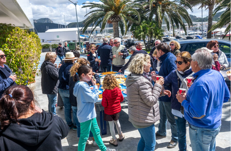 Grupo de personas en una reunión al aire libre junto a un muelle con yates, palmeras y montañas en el fondo.