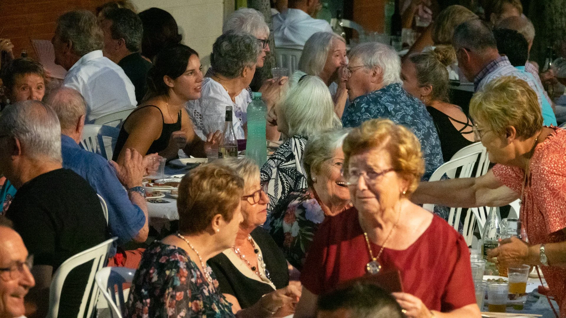 Grupo de personas mayores y jóvenes conversando y disfrutando en una reunión social, sentados en una mesa con comida y bebidas.