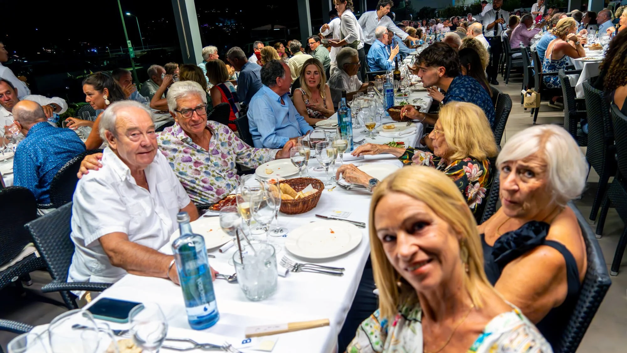 Grupo de personas en una cena formal en un restaurante, disfrutando de comida y bebidas, en un ambiente moderno y con muchas mesas y comensales.