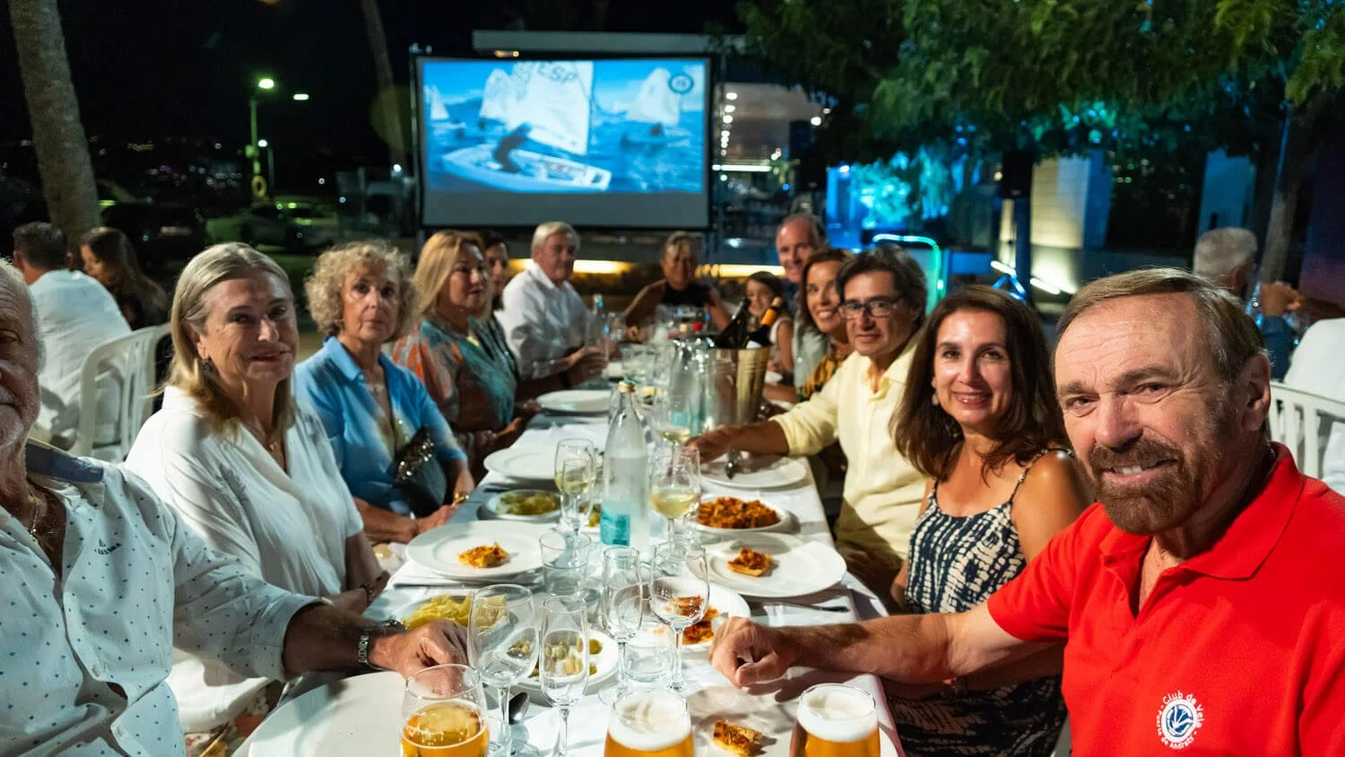 Grupo de personas sentadas en una mesa al aire libre durante la noche, disfrutando de comida y bebidas, con una pantalla grande en el fondo.