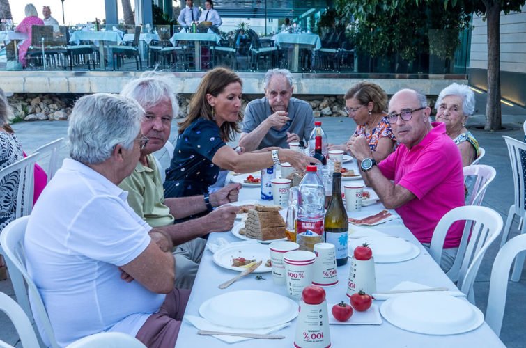 Personas mayores sentadas en una mesa en un restaurante al aire libre, compartiendo comida y bebidas, con decoraciones y otros comensales en el fondo.