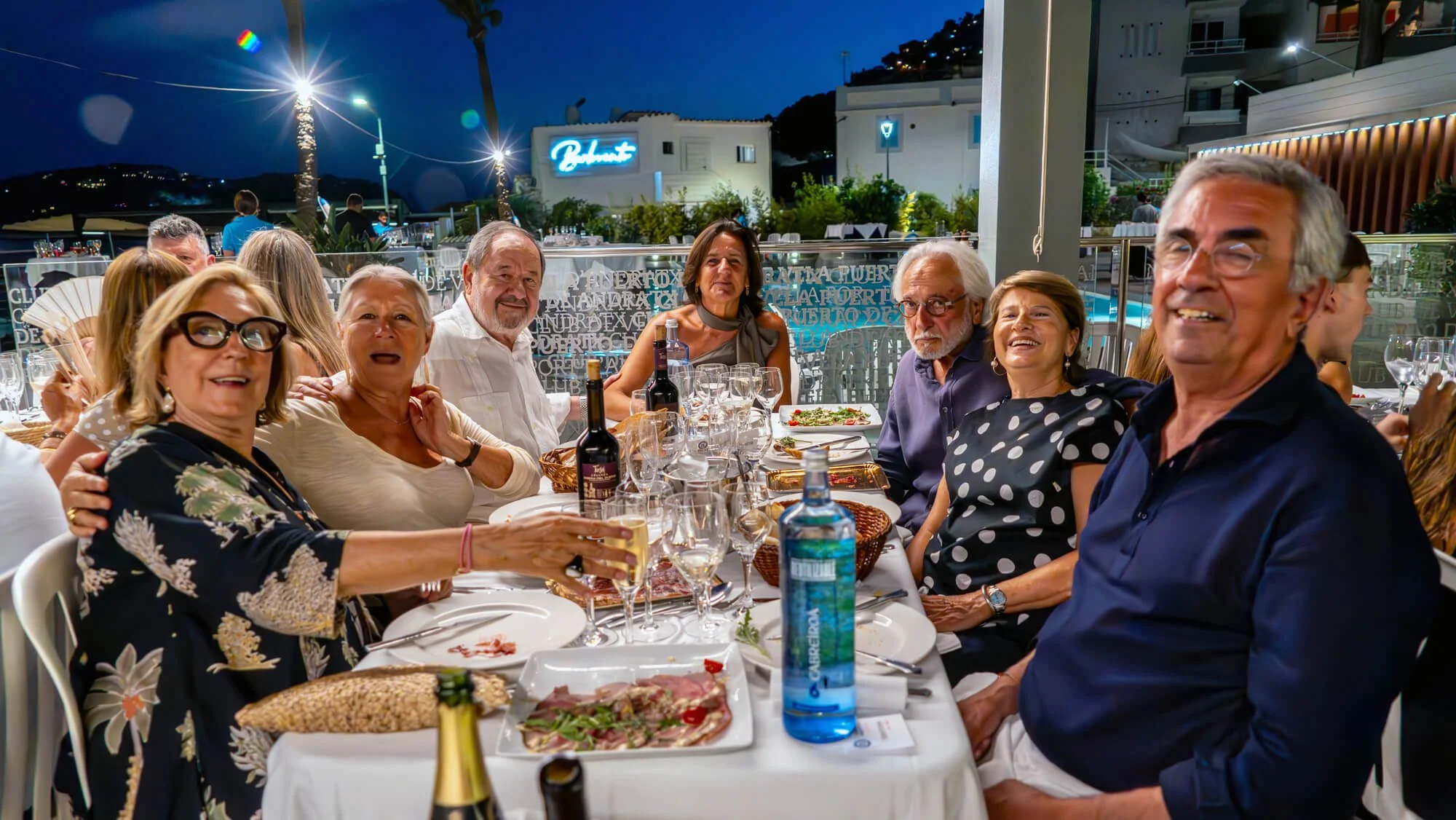 Grupo de personas riendo y disfrutando en una cena al aire libre en la noche, alrededor de una mesa con comida y vino, en un restaurante con decoración moderna y luces en el fondo.