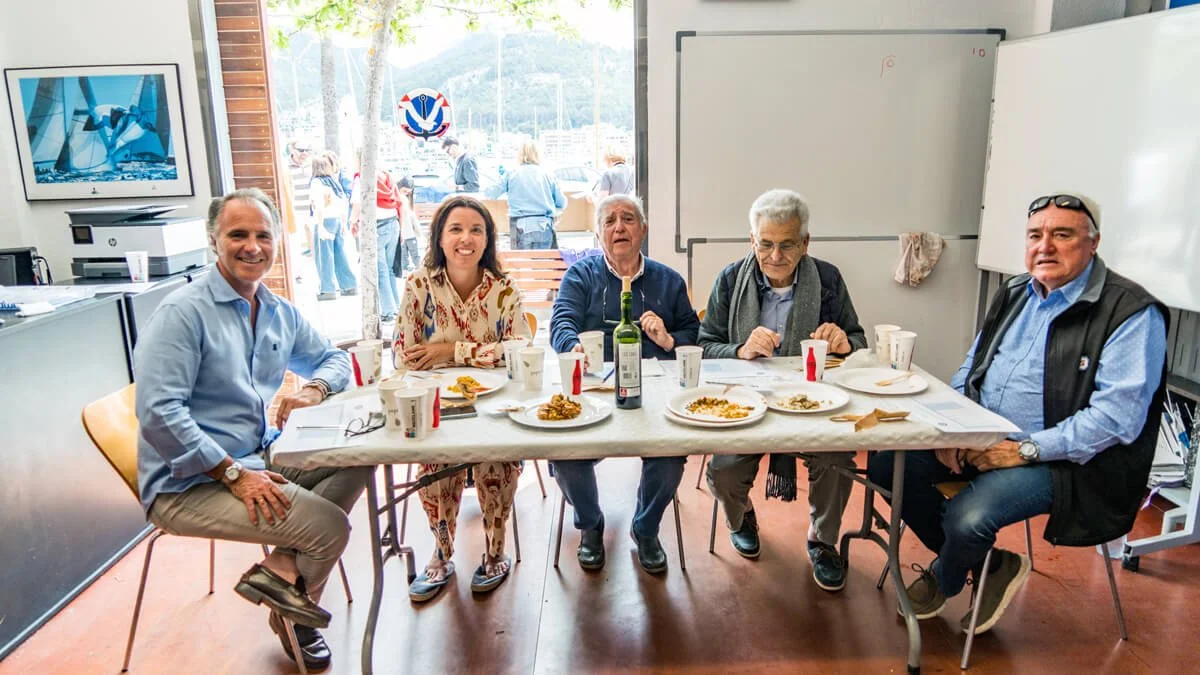 Seis personas sentadas en una mesa en una sala, riendo y comiendo, con comida y bebidas en platos y vasos, en un ambiente que parece un restaurante o cafetería.