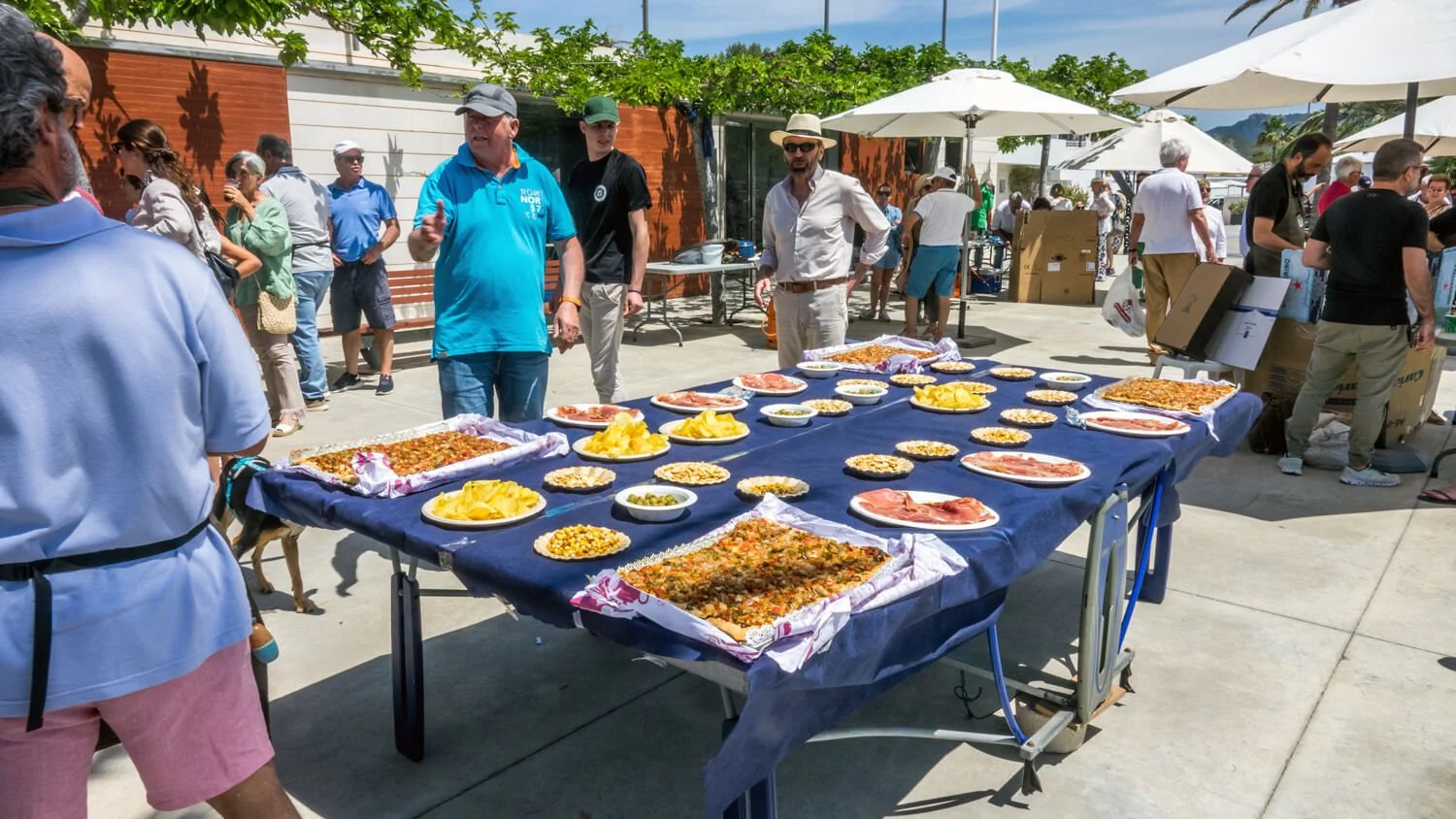 Mesa con comida en un evento al aire libre, con numerosas personas alrededor, algunas haciendo fila y otras conversando, en un día soleado con paraguas blancos y árboles en el fondo.