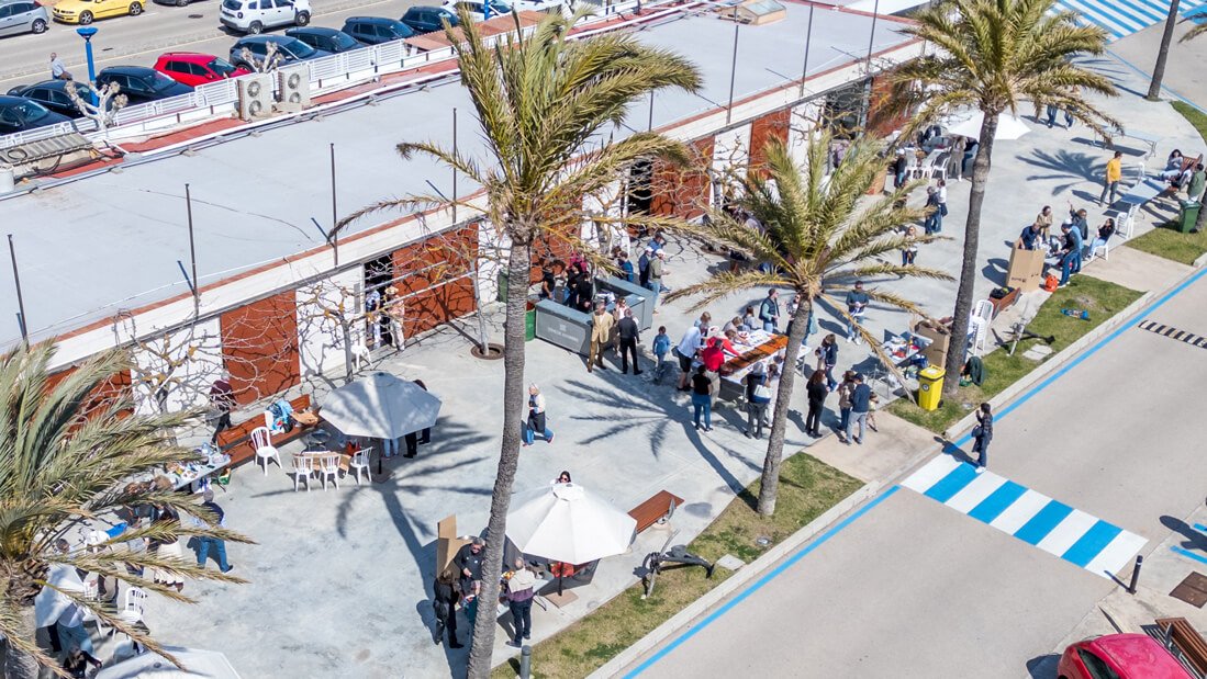 Escena de un evento al aire libre con muchas personas, mesas y sillas, palmeras y un edificio con paredes rojas y ventanas.