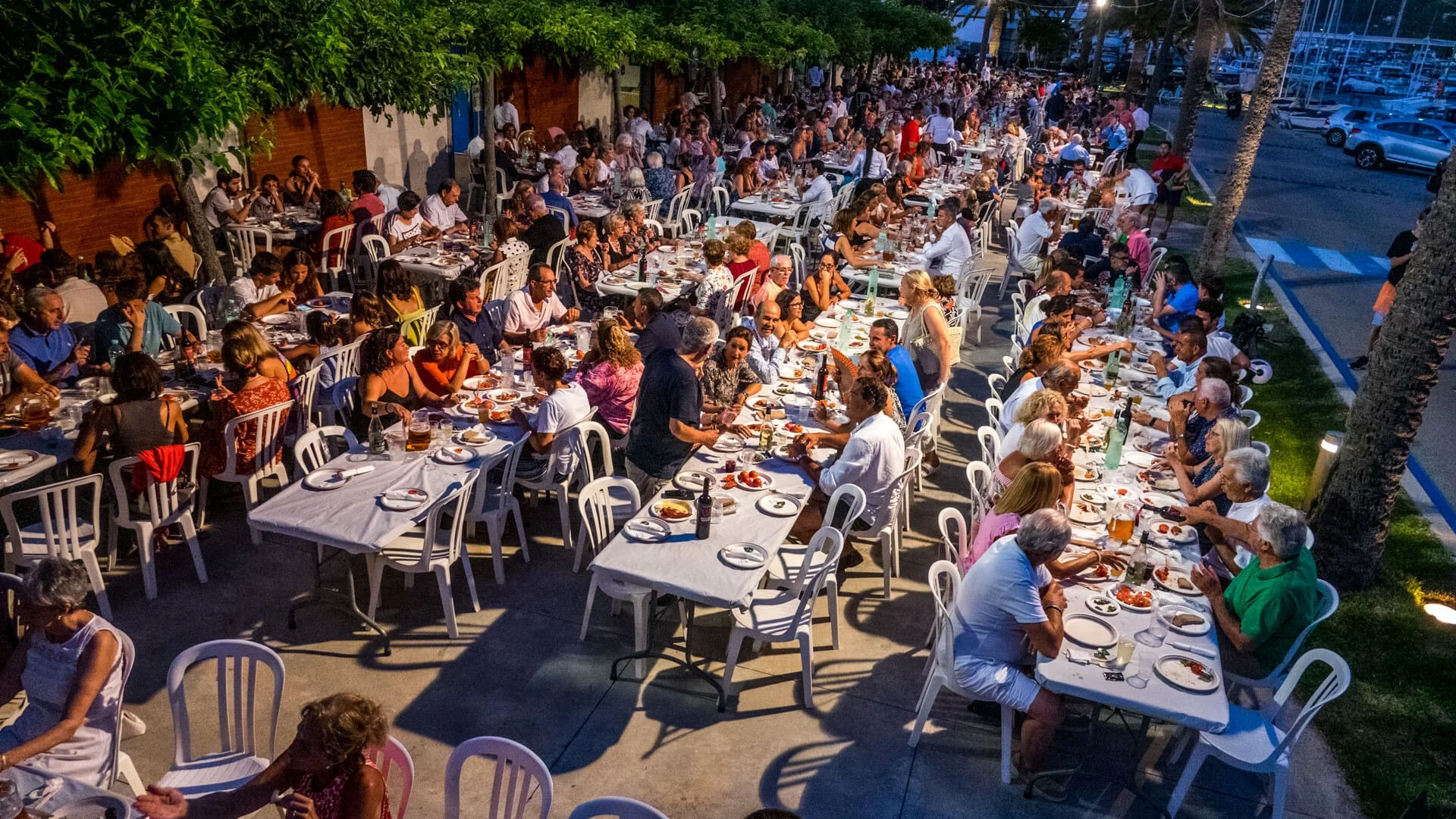 Una escena nocturna de una comida al aire libre con muchas personas sentadas en largas mesas, comiendo y socializando bajo árboles y luces de la calle.
