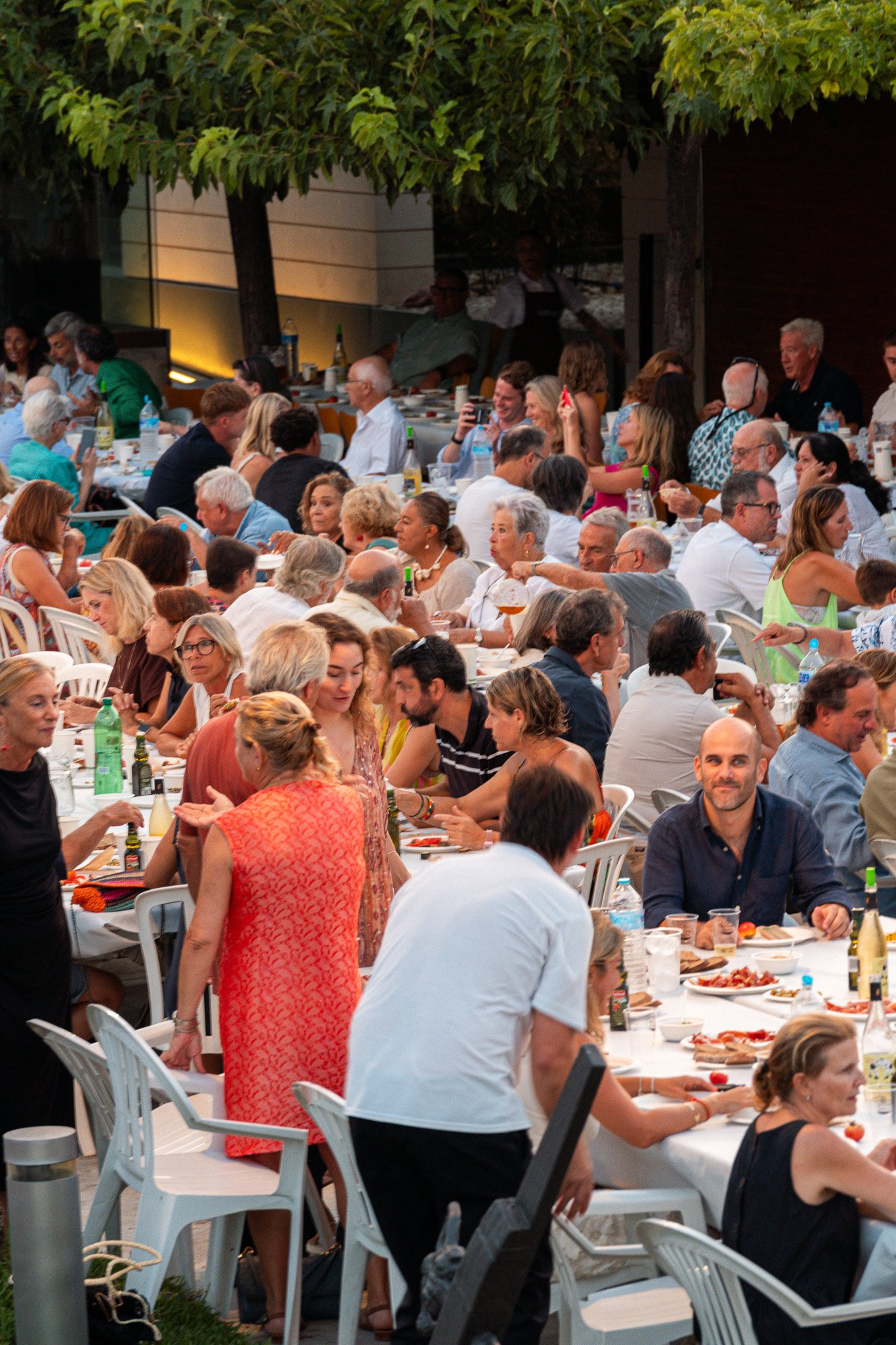 Un grupo de personas celebrando una comida al aire libre en una reunión social. La mesa está llena de comida, bebidas y utensilios, y las personas parecen estar disfrutando y conversando.