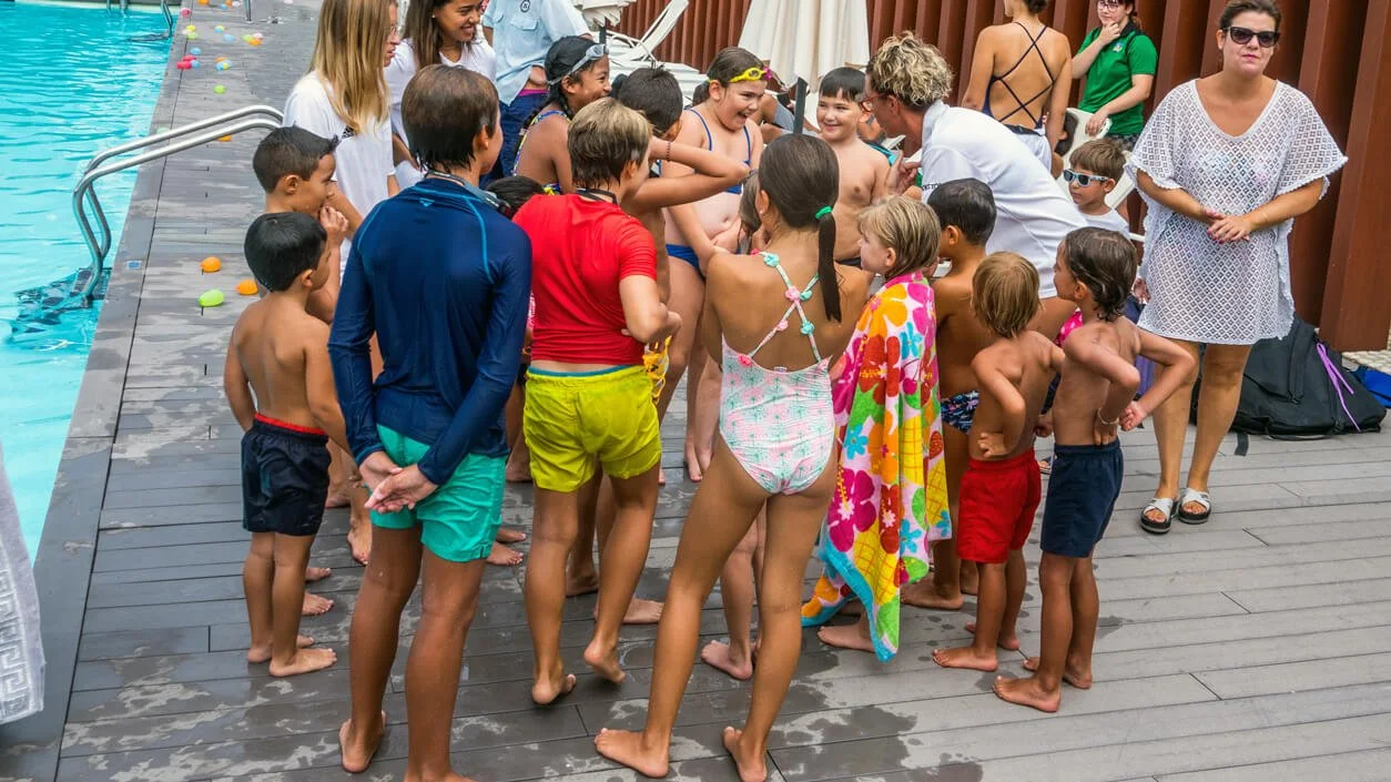 Niños y adultos en la piscina durante una actividad de natación, algunos con trajes de baño, algunos con ropa casual, y una mujer con gafas de sol.