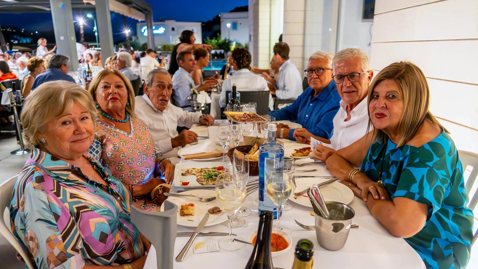 Grupo de personas mayores sentadas en una mesa en un restaurante al aire libre por la noche, disfrutando una comida juntos.