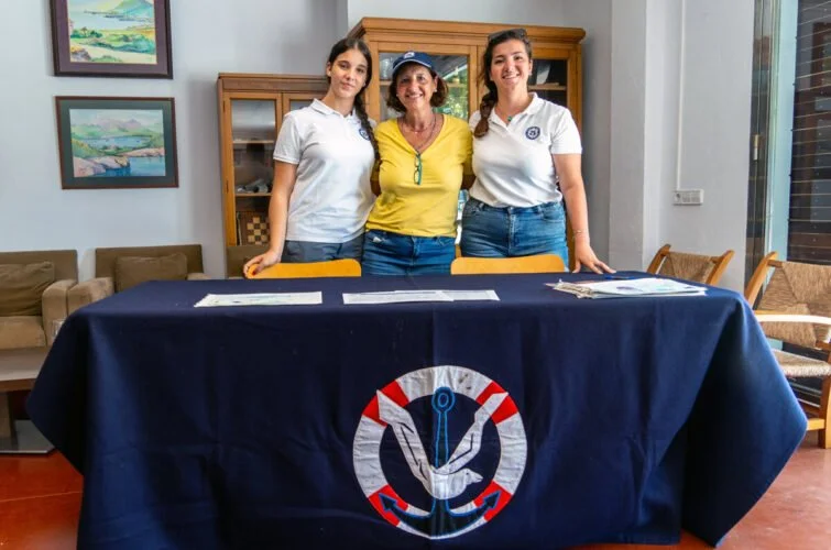 Tres mujeres de pie detrás de una mesa cubierta con una manta azul con un ancla dorada y rojo, en una sala con paredes decoradas y muebles de madera.