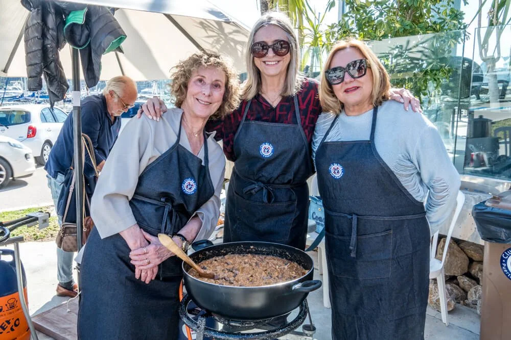 Tres mujeres con delantales negros cocinando y posando junto a una olla grande con comida en un toldo junto a un parque de automóviles y plantas.