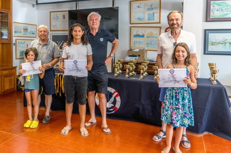 Niños y adultos en una ceremonia de premiación, con trofeos y certificados, en una sala con cuadros en las paredes.