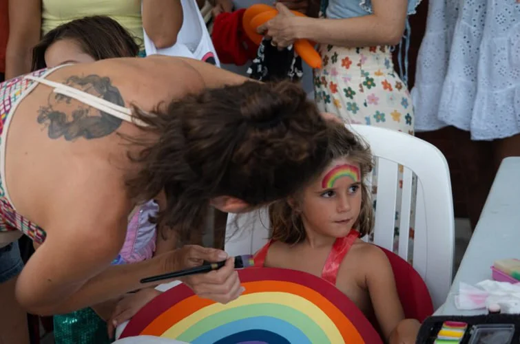 Niña con maquillaje de arcoíris en la cara, sentada en una silla con un dibujo de arcoíris, mientras una mujer le pinta otro arcoíris en la frente en un evento festivo.