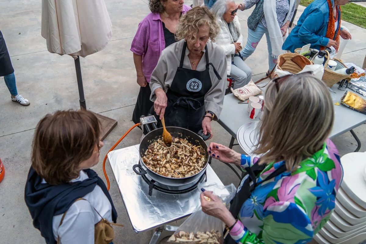 Mujer cocinando hongos en una sartén portátil, rodeada de varias personas en un evento al aire libre.