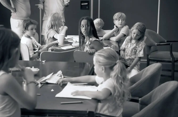 Niños y niñas sentados en una mesa, algunos con libros y otros con bebidas, en una sala de reuniones o aula.