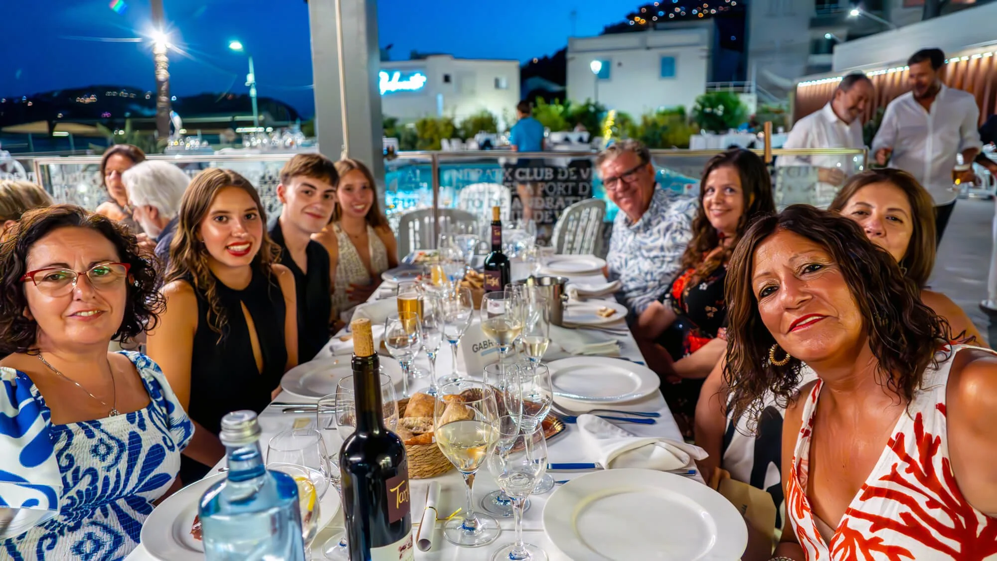 Grupo de personas celebrando en una cena al aire libre por la noche, con comida y bebidas en la mesa, en un ambiente festivo y alegre.