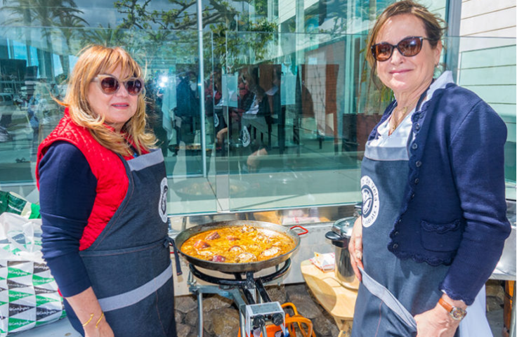 Dos mujeres cocinando en una calle con un sartén grande de comida, ambas usan delantales y gafas de sol, parece un evento al aire libre.