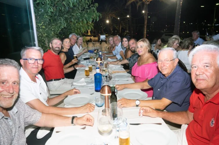 Un grupo de personas sentadas en una mesa larga en un restaurante al aire libre durante la noche, compartiendo una comida. Hay platos, bebidas y velas en la mesa.