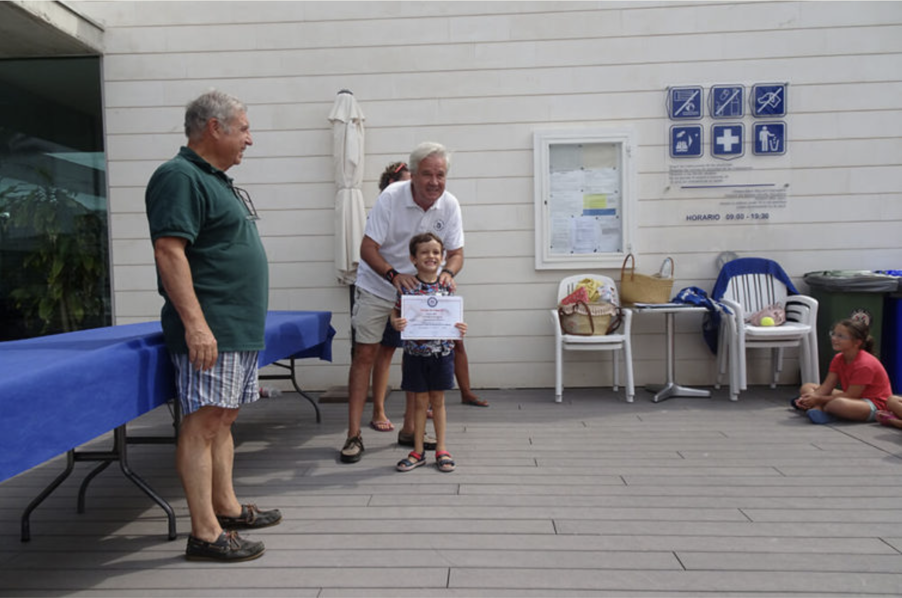 Un grupo de personas en una ceremonia de premiación en un espacio abierto. Un niño recibe un certificado, una mujer lo acompaña y dos hombres observan, mientras otras personas están sentadas a un lado.