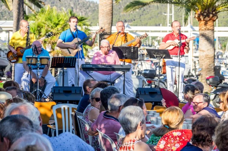 Un grupo de músicos tocando en vivo en un escenario al aire libre, con una audiencia sentada disfrutando la actuación.