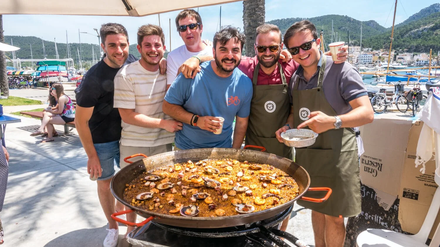 Grupo de amigos disfrutando de una comida al aire libre en un muelle, con mar y barcos de fondo.