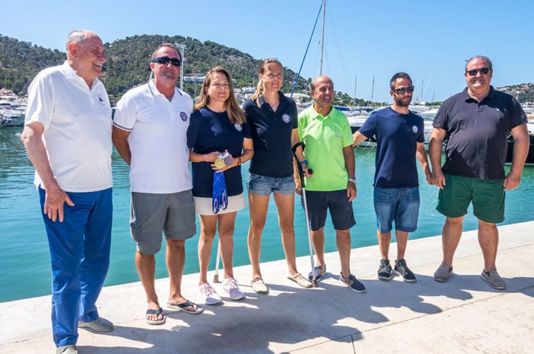 Grupo de siete personas posando junto a un muelle con agua y yates, en un día soleado con montañas al fondo.