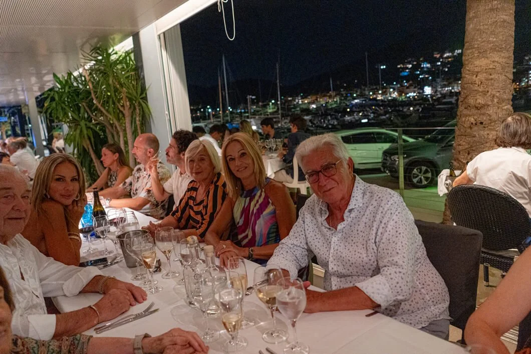 Grupo de personas disfrutando de una cena en un restaurante en la noche, con vista a un puerto con yates y barcos.
