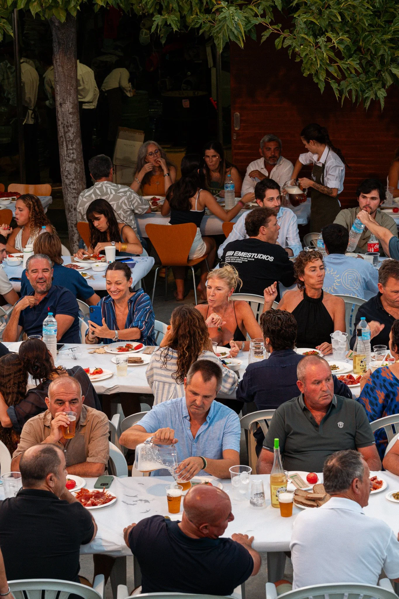 Un grupo de personas sentadas en mesas redondas disfrutando de comida y bebidas en una evento social, con varias mesas llenas de comida, bebidas y platos, en un entorno al aire libre con árboles y paredes de madera.