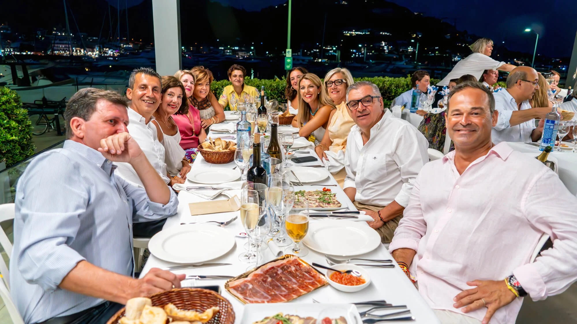 Grupo de personas en una cena al aire libre en la noche, en una mesa con comida, bebidas y vino, con fondo de un puerto con yates