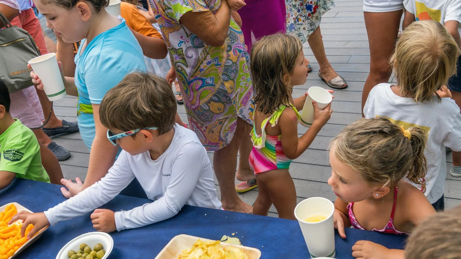 Niños y niñas en una fiesta al aire libre, algunos con vasos de bebida, otros sirviéndose comida en una mesa. Todos parecen estar disfrutando un evento social en un lugar público con piso de madera.