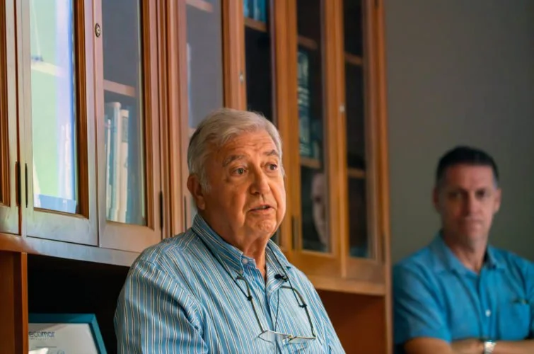 Dos hombres en una sala de reuniones, uno en primer plano y otro en segundo plano, con estanterías de libros de fondo.